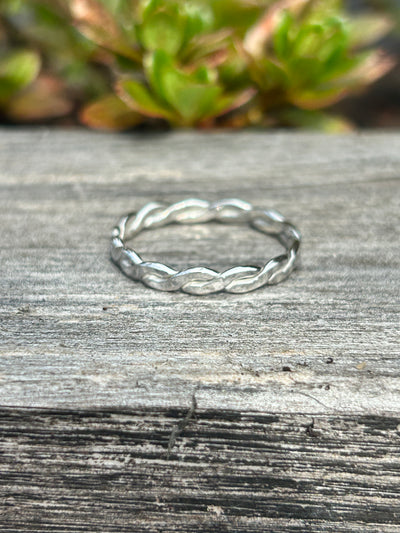 Silver braided stacking ring on a wooden surface with greenery in the background