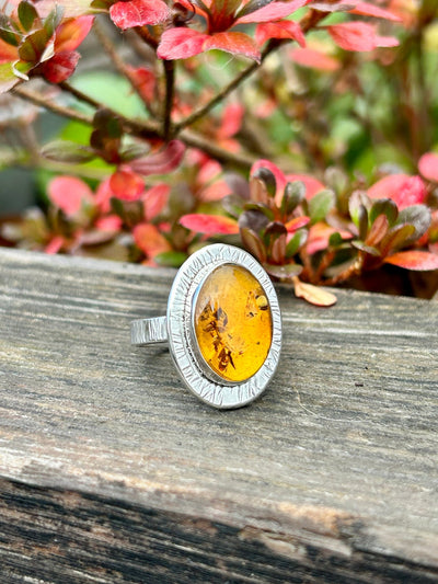 Silver ring with amber stone on a wooden surface with blurred red and green foliage in the background