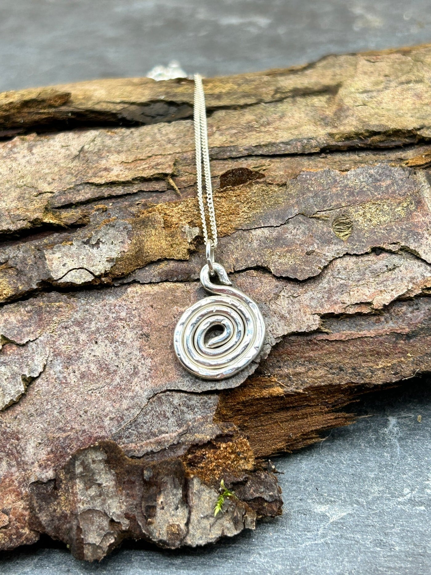 A silver pendant in the shape of a small spiral, attached to a fine chain, displayed on a rustic wooden surface.