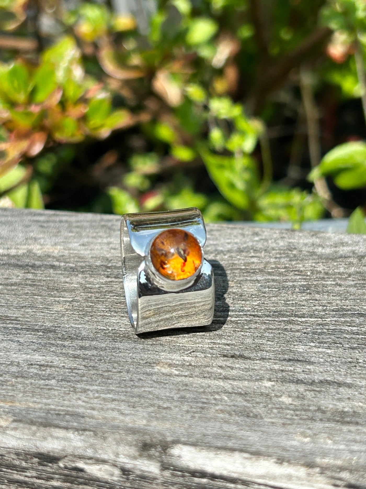 wide sterling silver band with an amber cabochon by LaVidaLoca Jewellery shown on a wooden background with green foliage